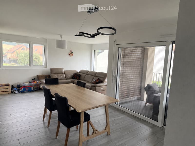 Bright living and dining area with a beige sofa, wooden dining table, black chairs, and a large window with blinds.