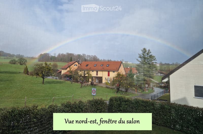 A two-story house with a red-tiled roof and beige walls, situated in a green, hilly landscape with a visible rainbow in the background.