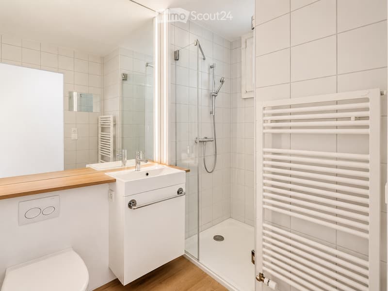 Modern bathroom with white tiles, a glass-enclosed shower, a sink with a wooden countertop, a toilet, and a white radiator.