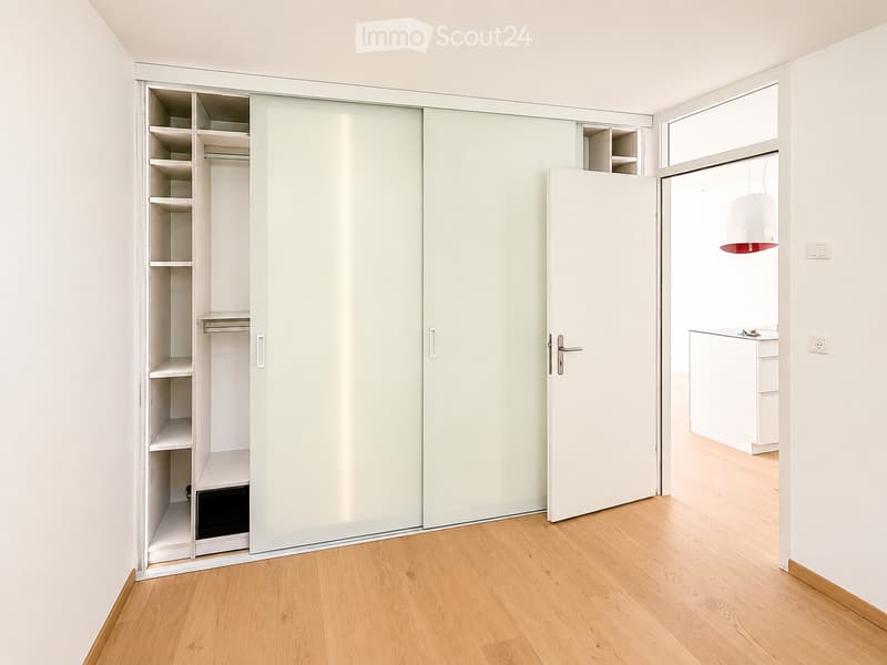 Empty room with light-colored wooden flooring, white walls, and a large sliding closet with open shelving. A doorway leads to another room with a visible kitchen area.