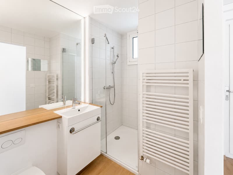 Modern bathroom with white tiles, wooden countertop, sink, mirror, shower, radiator, and toilet.