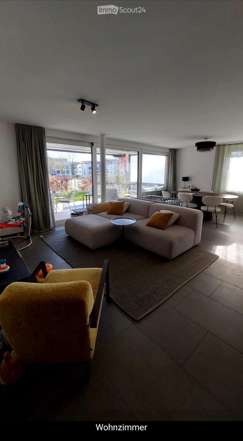 Modern living room with a large sectional sofa, coffee table, and dining area with chairs and a table. Large windows and sliding doors lead to a balcony or outdoor area.