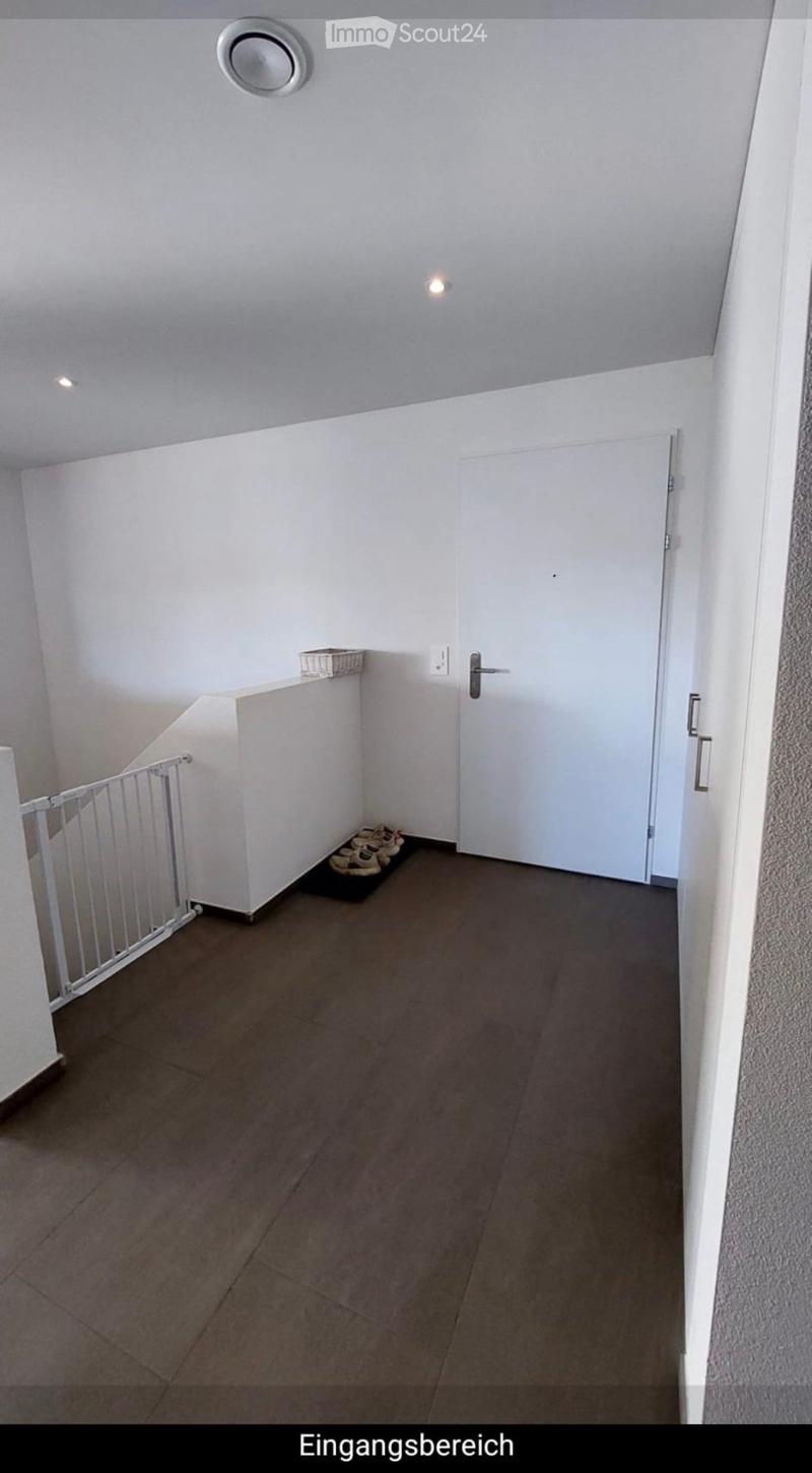 Empty hallway with white walls, gray tiled floor, a white door, a small shelf with a basket, and a few pairs of shoes placed near the wall.