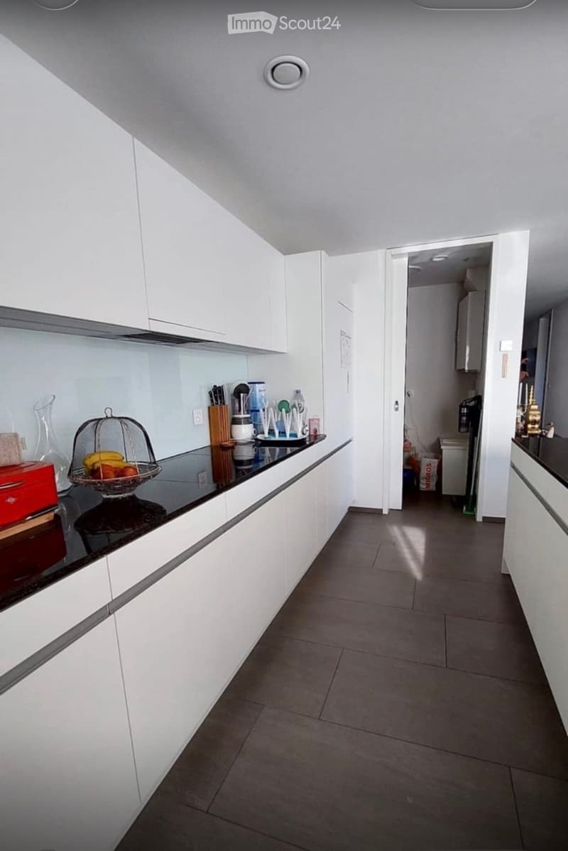 Modern kitchen with white cabinets, dark countertop, fruit basket, knife block, and a doorway leading to another room.