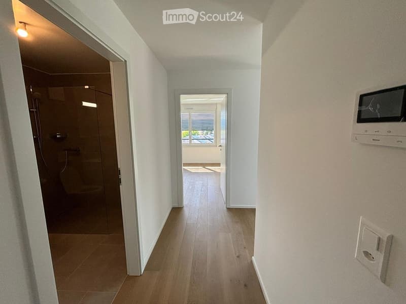 Empty hallway with wooden flooring, white walls, and doors leading to a bathroom and another room with a view.