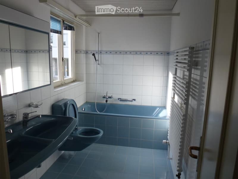 Bathroom with blue tiles, a sink, toilet, bathtub, and a showerhead. Features a window with blinds and a radiator.