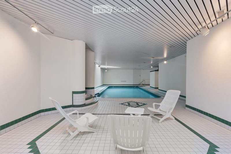 Indoor pool area with white lounge chairs, small table, and tiled flooring.