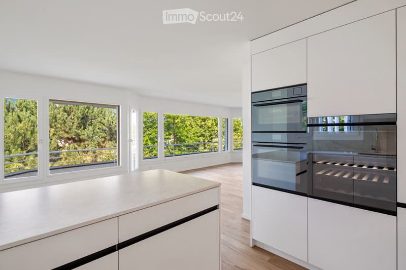 Modern kitchen with white cabinets, a central island, and large windows offering a view of greenery outside.