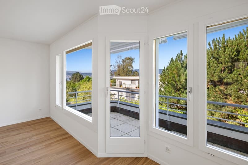 Empty room with wooden flooring, large windows, and a view of trees and a distant landscape.