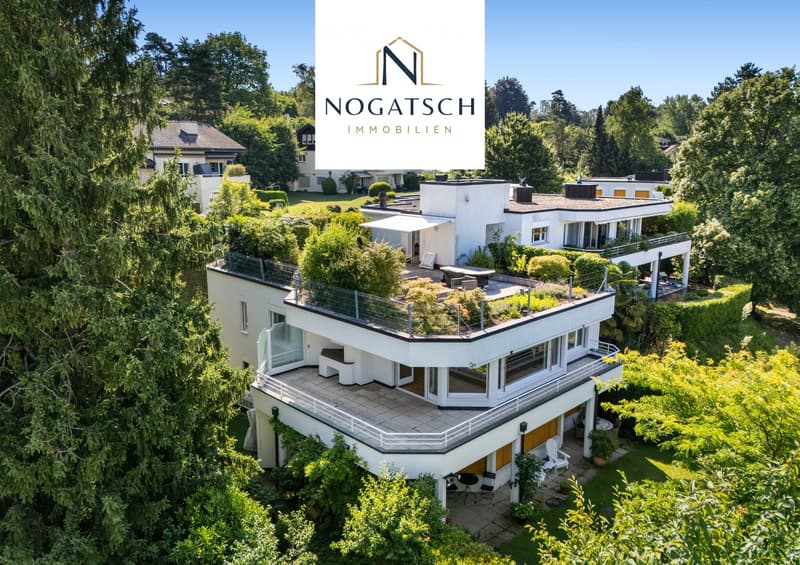 Aerial view of a modern, white multi-story house with large windows and balconies, surrounded by lush greenery and trees. The house features a flat roof and is part of a residential area with other houses visible in the background.
