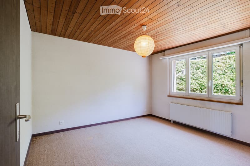 Empty room with light-colored walls, wooden ceiling, carpeted floor, large windows, radiator, and a hanging paper lantern light fixture.