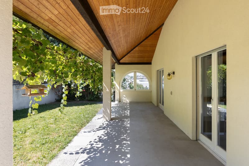 A covered outdoor corridor with a wooden ceiling, concrete floor, and glass doors leading to a grassy area with a bird feeder.