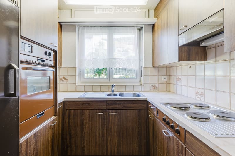 A well-equipped kitchen with wooden cabinets, a stainless steel sink, a four-burner electric stove, and a built-in oven. The space features tiled backsplash and a window with white curtains.