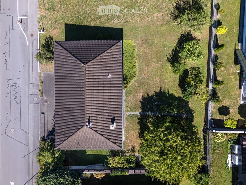 Aerial view of a two-story house with a dark roof, surrounded by a grassy yard and trees. The house has a chimney and is situated near a road with visible markings.
