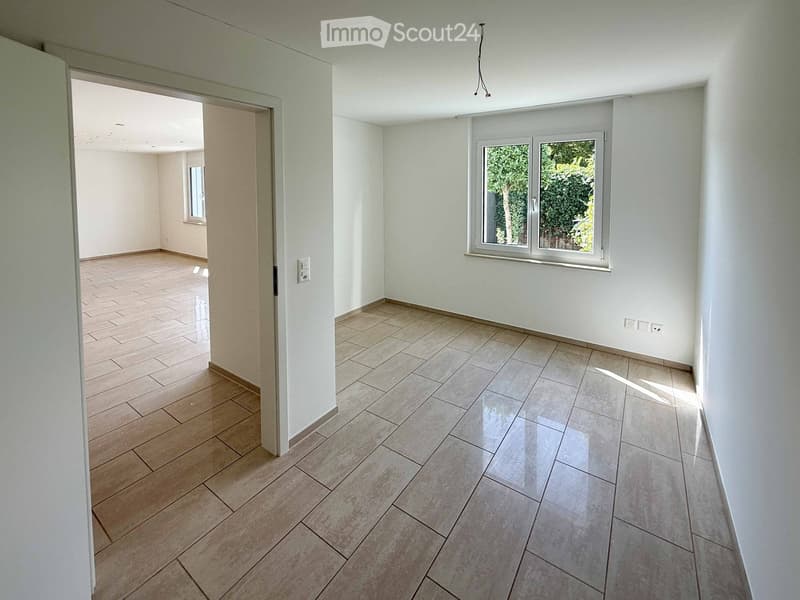 Empty room with tiled floor and white walls, featuring a large window with a view of greenery and an open doorway leading to another room.