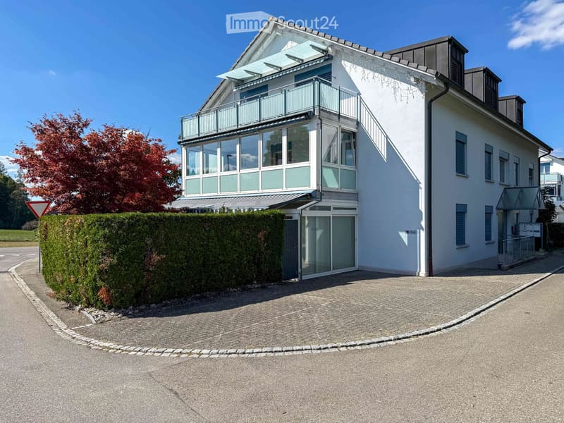 Modern two-story house with white exterior, large windows, and a balcony on the upper level. The house features a well-maintained hedge and a driveway.