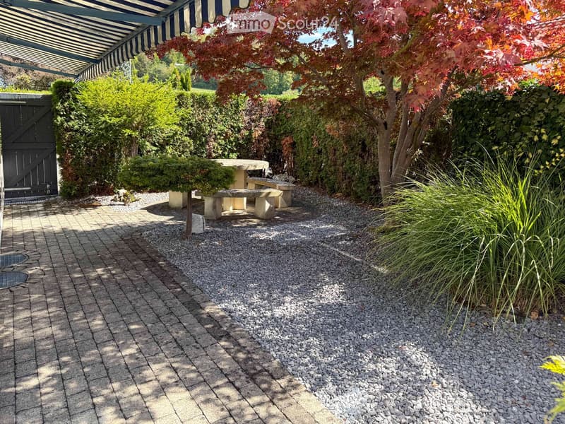 Well-maintained garden with a stone table and benches, surrounded by various plants and trees, including a red-leaved tree, under a striped canopy.