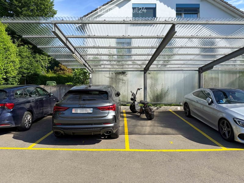 A modern covered parking area with three cars and a scooter parked under a transparent roof structure, with a white building in the background.