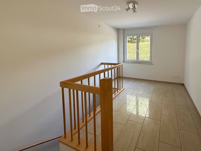 Empty hallway with tiled floor, white walls, wooden railing, and a window showing greenery outside.