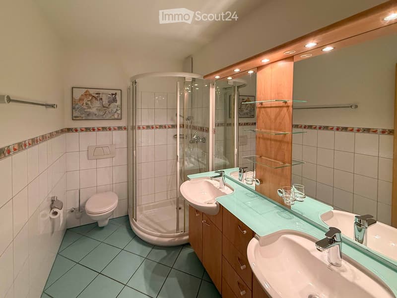 Modern bathroom with two sinks, a toilet, a glass-enclosed shower, and a large mirror with built-in lighting. The space features light blue tiles on the floor and white tiles on the walls, complemented by a decorative border and a framed picture.