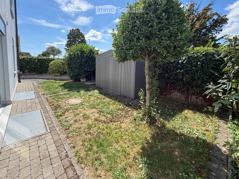 Garden area with a paved pathway, small grassy patch, trees, and a shed.