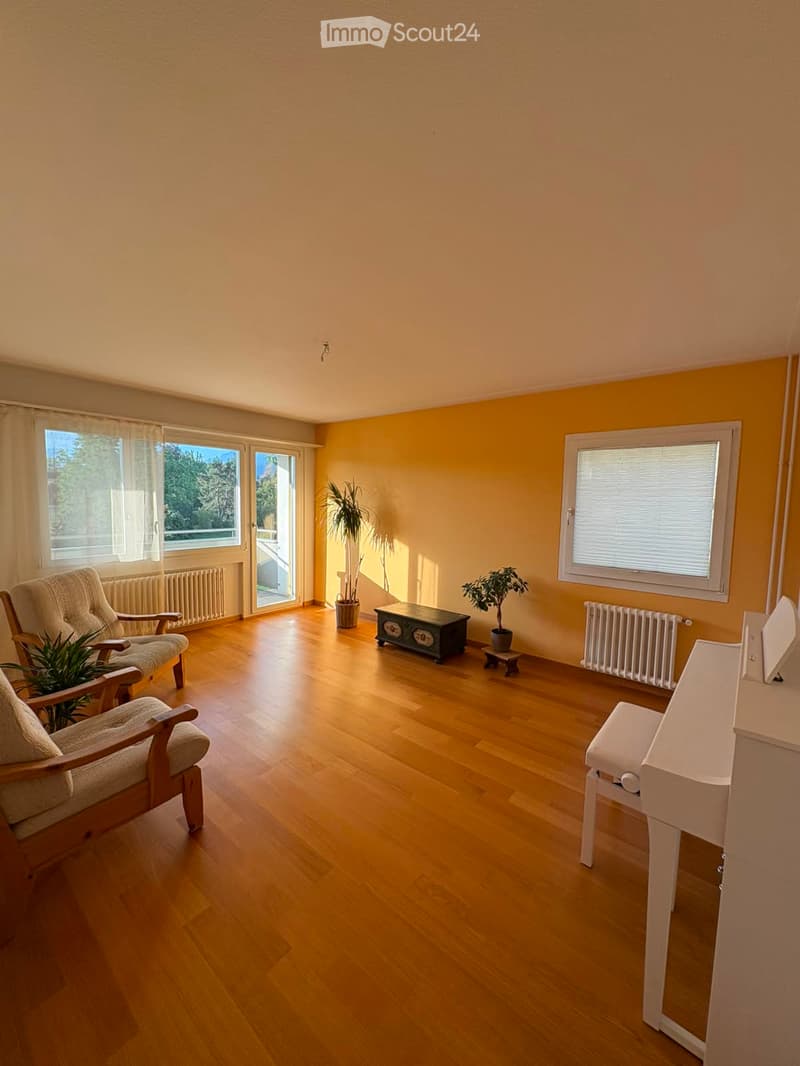 Bright living room with wooden flooring, large windows, and a sliding glass door leading to a balcony. Features include a sofa, a small table, two armchairs, and potted plants.