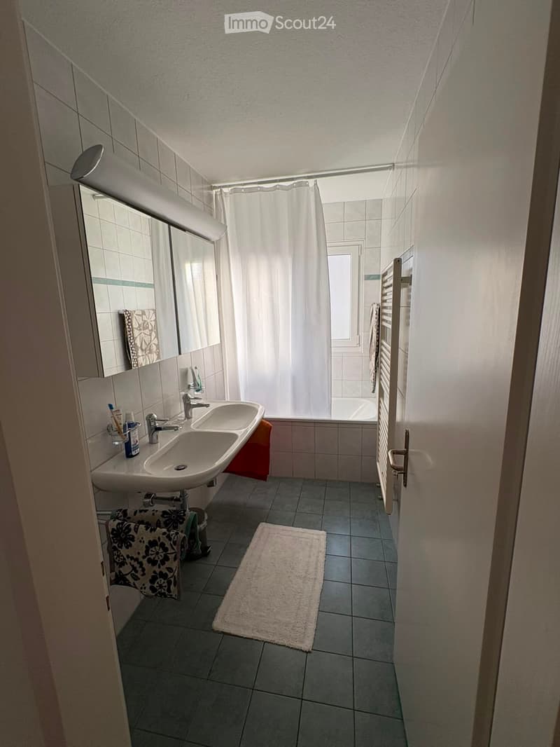 Modern bathroom with double sink, mirror, toothbrush holder, towels, bathrobe, and a shower area with a curtain. The floor is tiled, and there is a white rug in front of the sink.