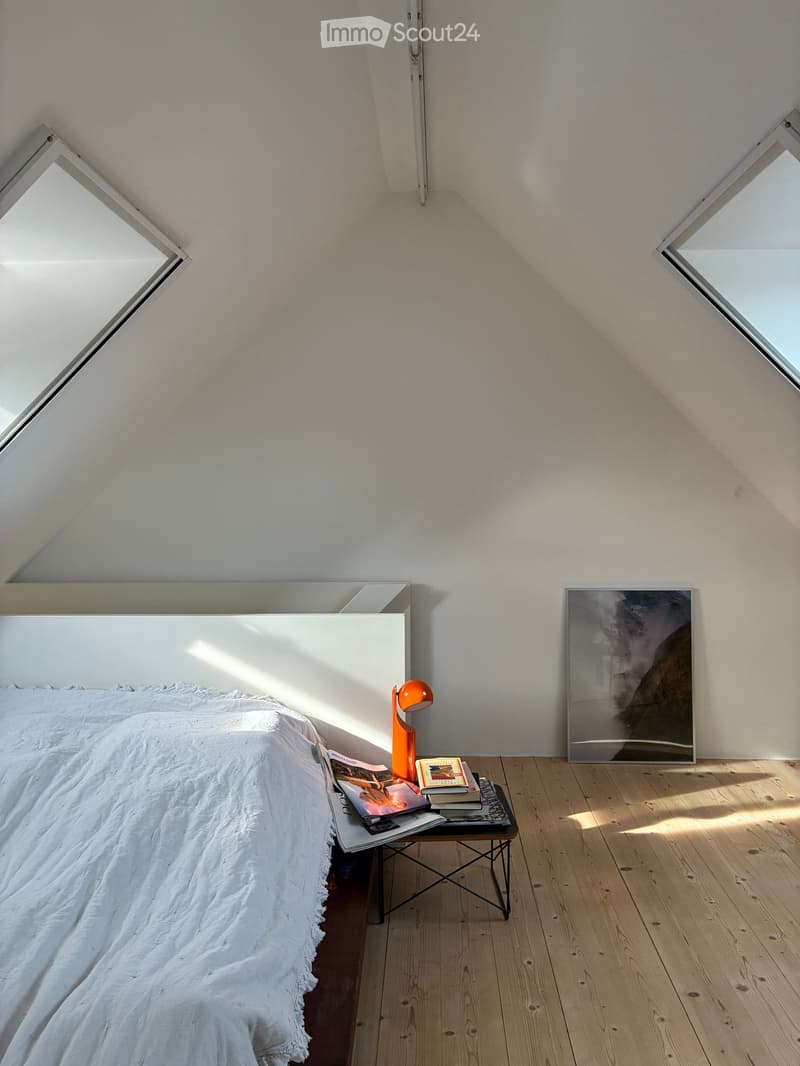 Bright, minimalist bedroom with wooden floors, white walls, and skylights. Features a white bed with a textured duvet, a small black nightstand with books and a lamp, and a large abstract picture frame on the wall.
