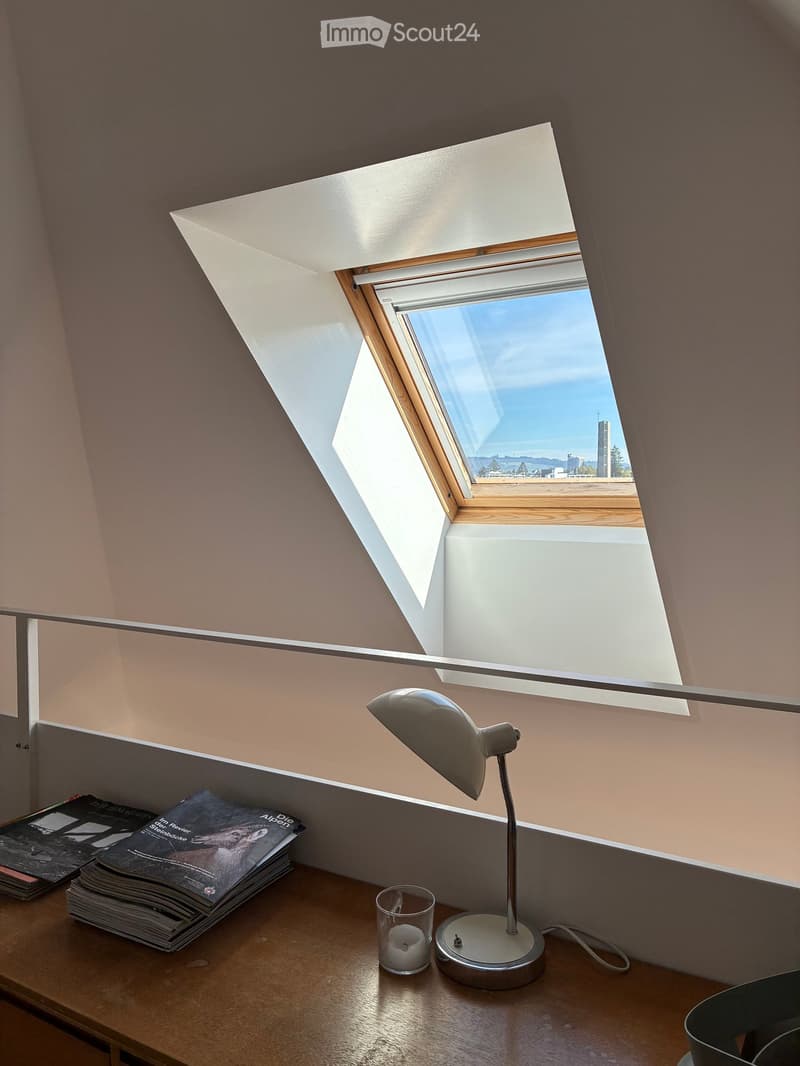 A cozy attic room with a slanted ceiling and skylight window, featuring a wooden table with a lamp, magazines, and a candle.