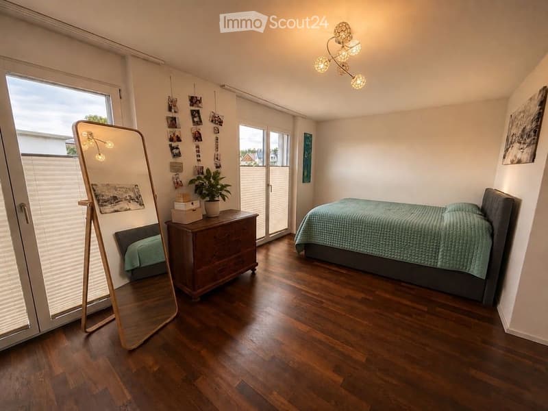 Cozy bedroom with wooden flooring, a neatly made bed, a nightstand with a plant, a large mirror, and several picture frames on the wall.