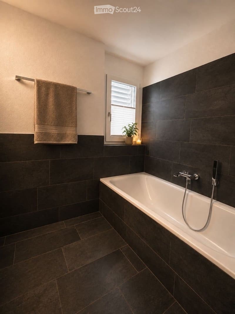 Modern bathroom with dark tiled walls and floor, a white bathtub, a towel on a rack, a small plant on the windowsill, and a candle.
