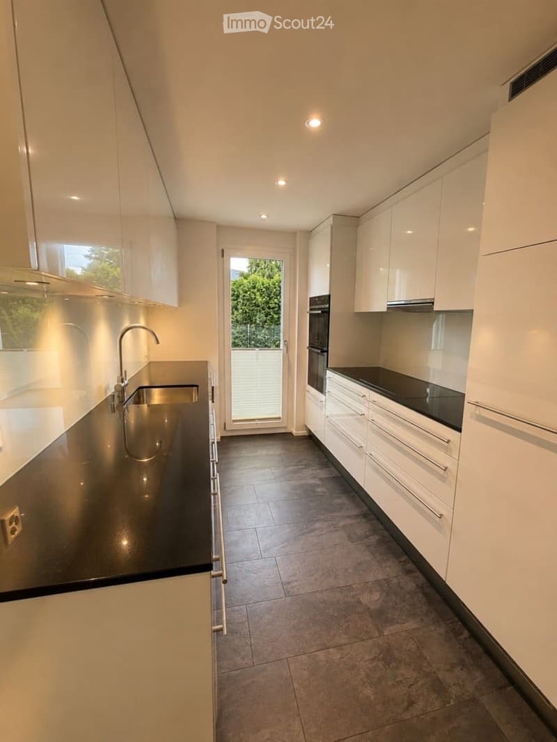 Modern kitchen with white cabinets, black countertops, stainless steel sink, and built-in oven. Features recessed lighting and a large window with blinds.