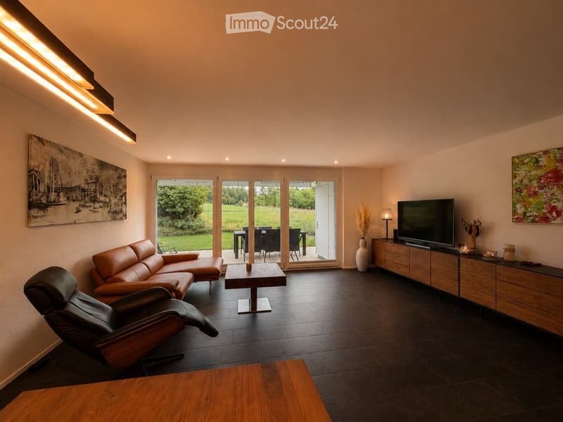 Modern living room with dark tiled flooring, large glass doors opening to a green outdoor area, leather sofa, coffee table, TV stand, and decorative vases.