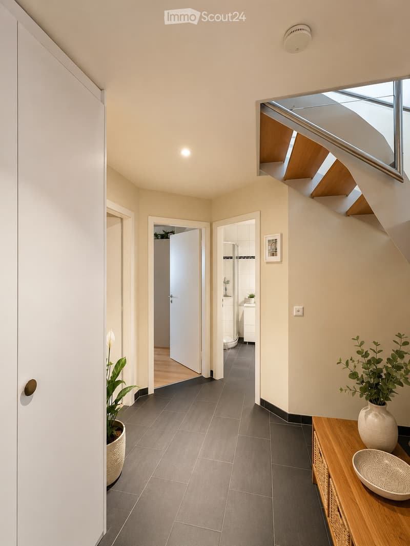 Modern hallway with grey tiled floor, white walls, and recessed lighting. Features a small wooden console table with a decorative bowl and a potted plant, leading to a bathroom and another room.