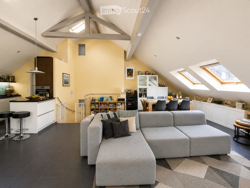 Modern, open-concept living and dining area with a grey sectional sofa, black stools, a dining table with chairs, a kitchen island, and built-in shelving. The space features exposed wooden beams and skylights.