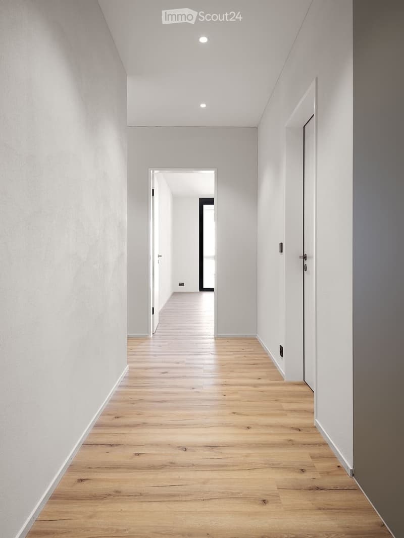 A modern, minimalist hallway with light wooden flooring, white walls, and recessed ceiling lighting. The hallway features several doors and a visible electrical outlet on the right wall.