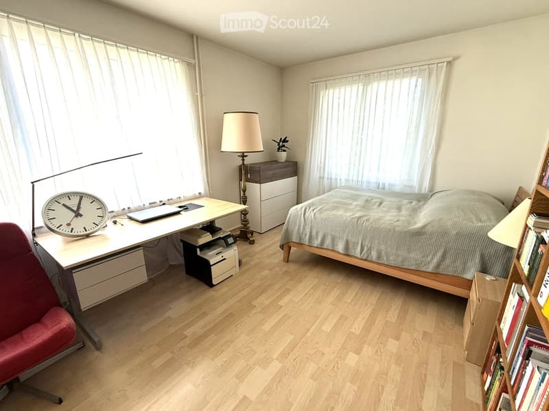 Cozy bedroom with a wooden bed, desk, chair, and bookshelf. Features a large window with sheer curtains, a nightstand with a lamp, and a small plant.