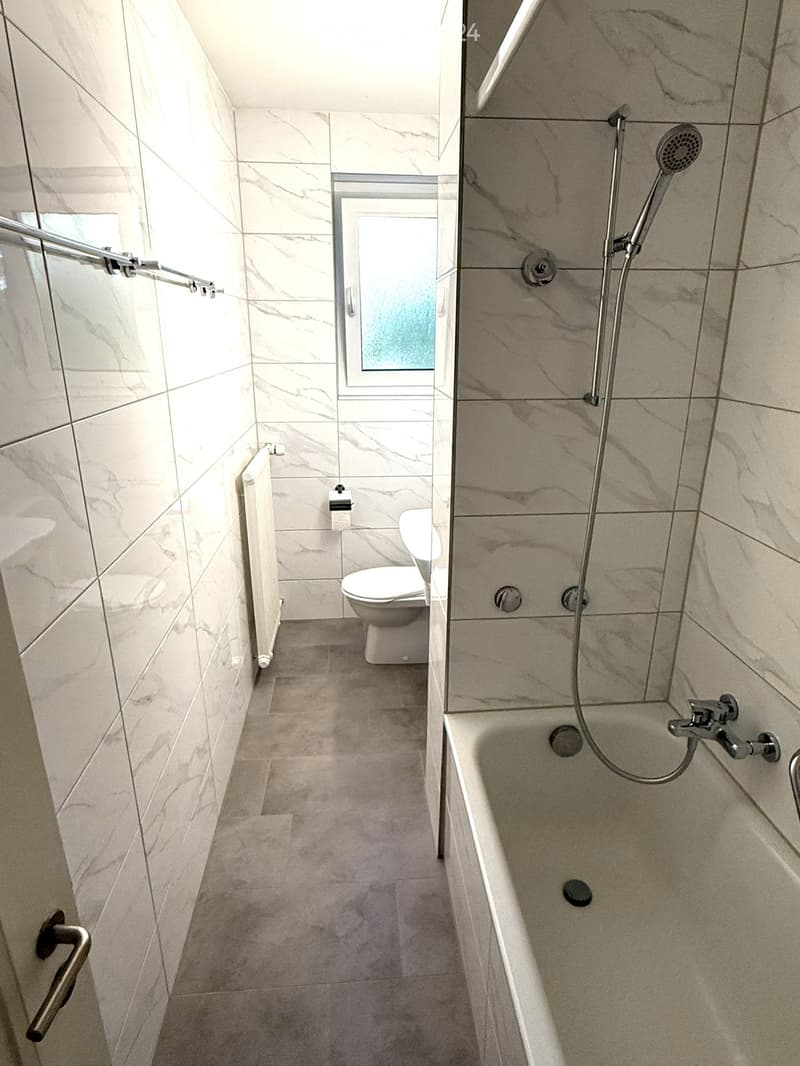 Modern bathroom with marble tiles, bathtub, shower, toilet, radiator, and window.