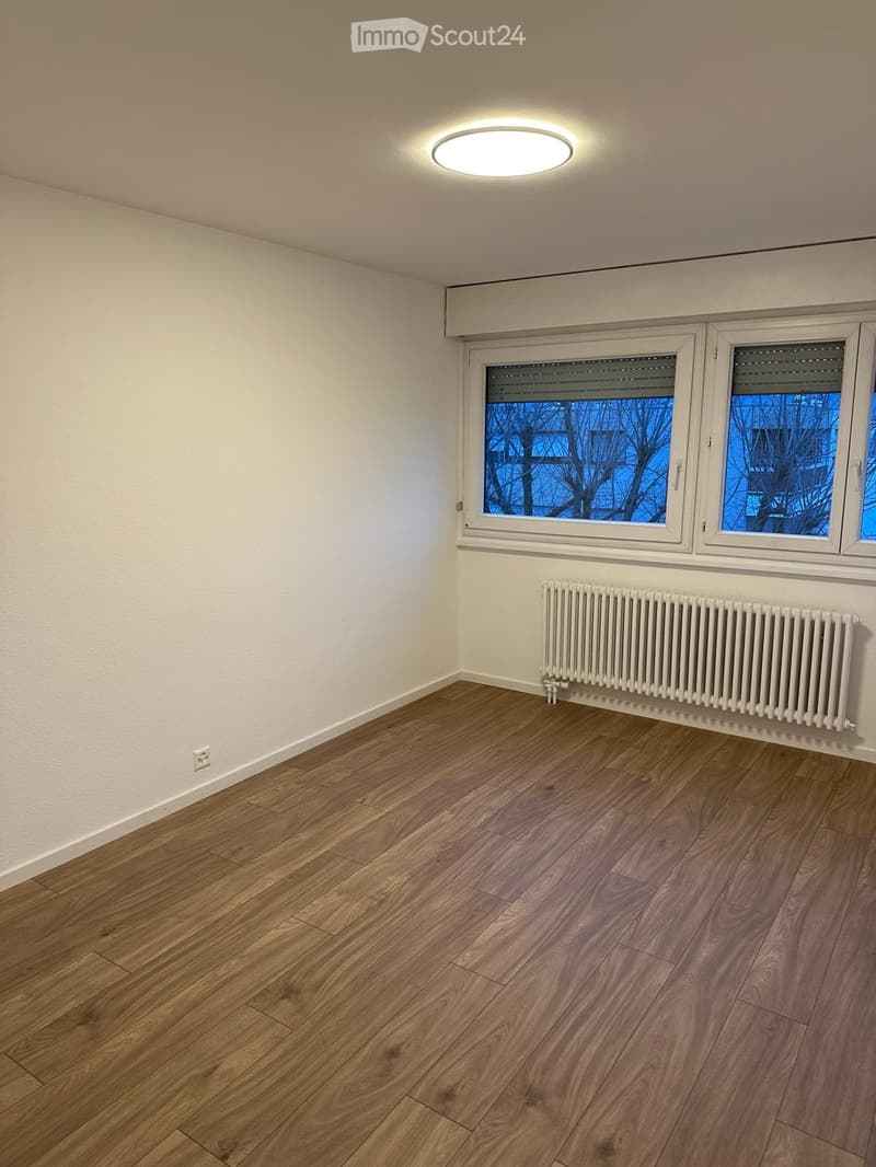Empty room with wooden flooring, white walls, large windows, and a radiator. No furniture or equipment present.