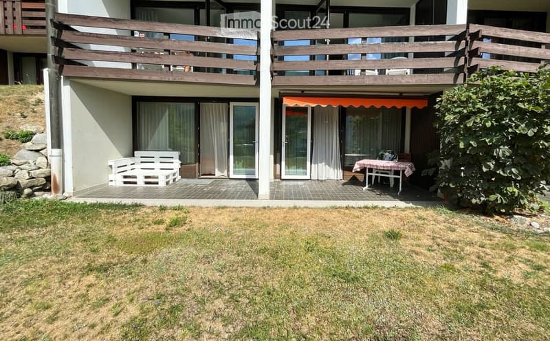 Outdoor patio area with a wooden bench, a small table with chairs, and an orange awning. The patio is connected to a house with large windows and a wooden balcony above.
