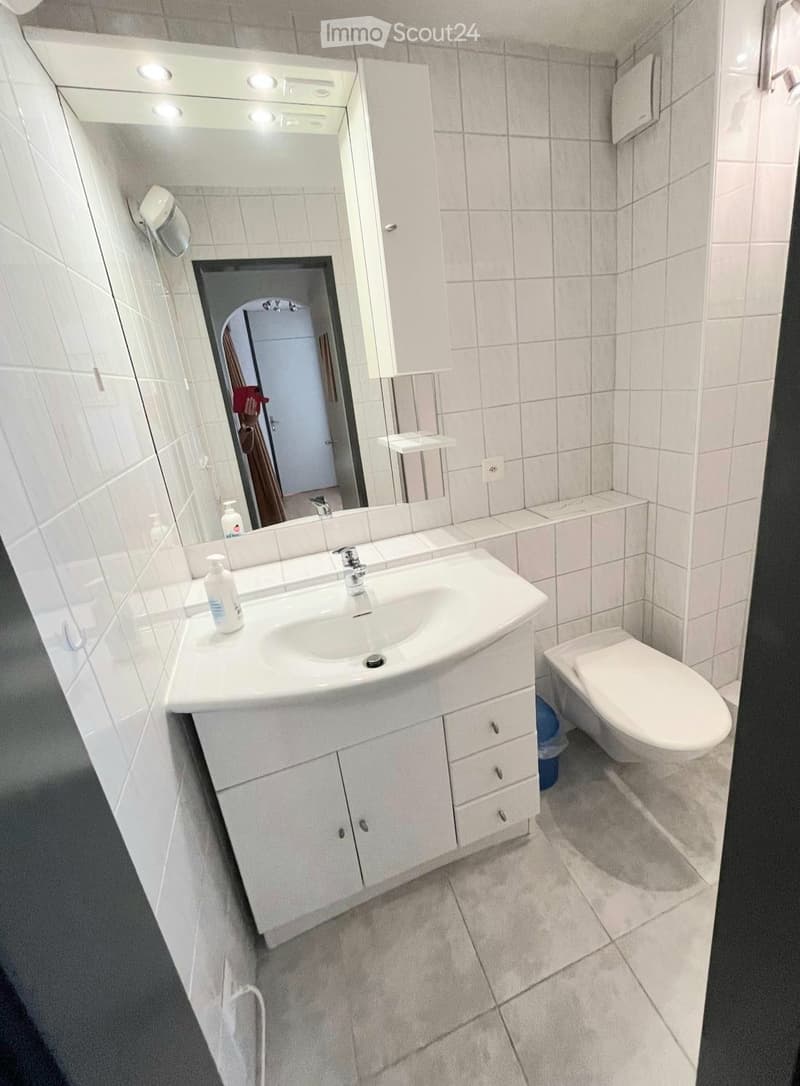 Modern bathroom with white tiled walls and floor, a white sink with a cabinet below, a toilet, and a large mirror with built-in lighting above the sink.