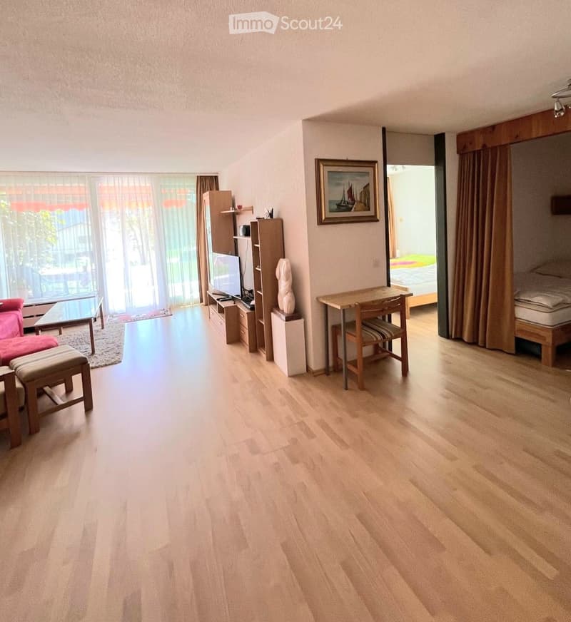 A modern, well-lit living space with light wooden flooring, a red sofa with an ottoman, a coffee table, a TV on a shelf, a small dining area with a table and chairs, and a bedroom area with a bed and curtains.