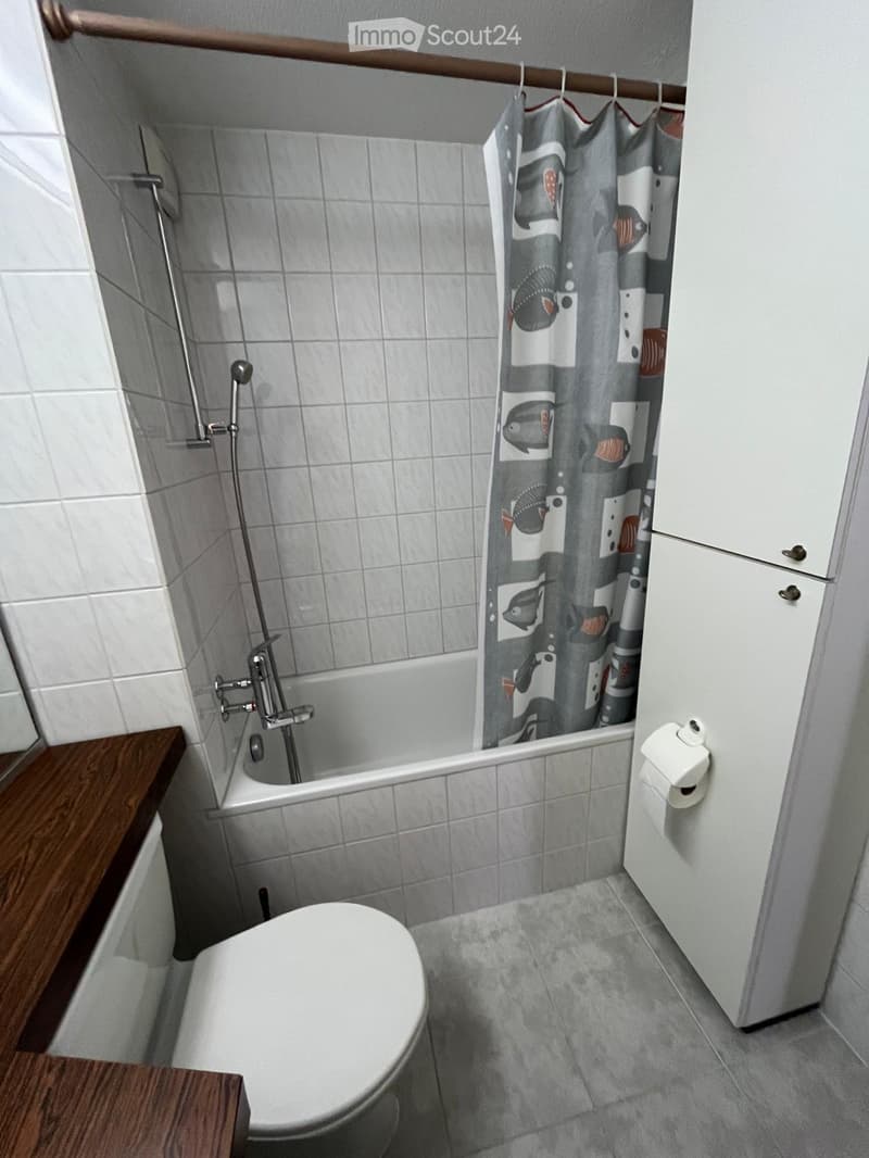 Small bathroom with a toilet, bathtub, shower curtain, and a wooden shelf.