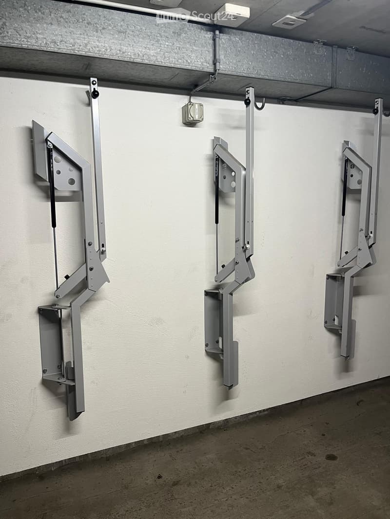 A concrete-floored hallway with three wall-mounted metal brackets or holders, each featuring a rectangular, open-fronted compartment with visible screws and bolts.