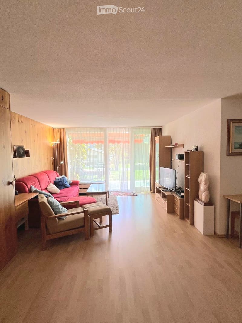 Bright living room with a red sofa, wooden chair, ottoman, coffee table, TV, shelves, and a statue. Large windows with curtains provide natural light.