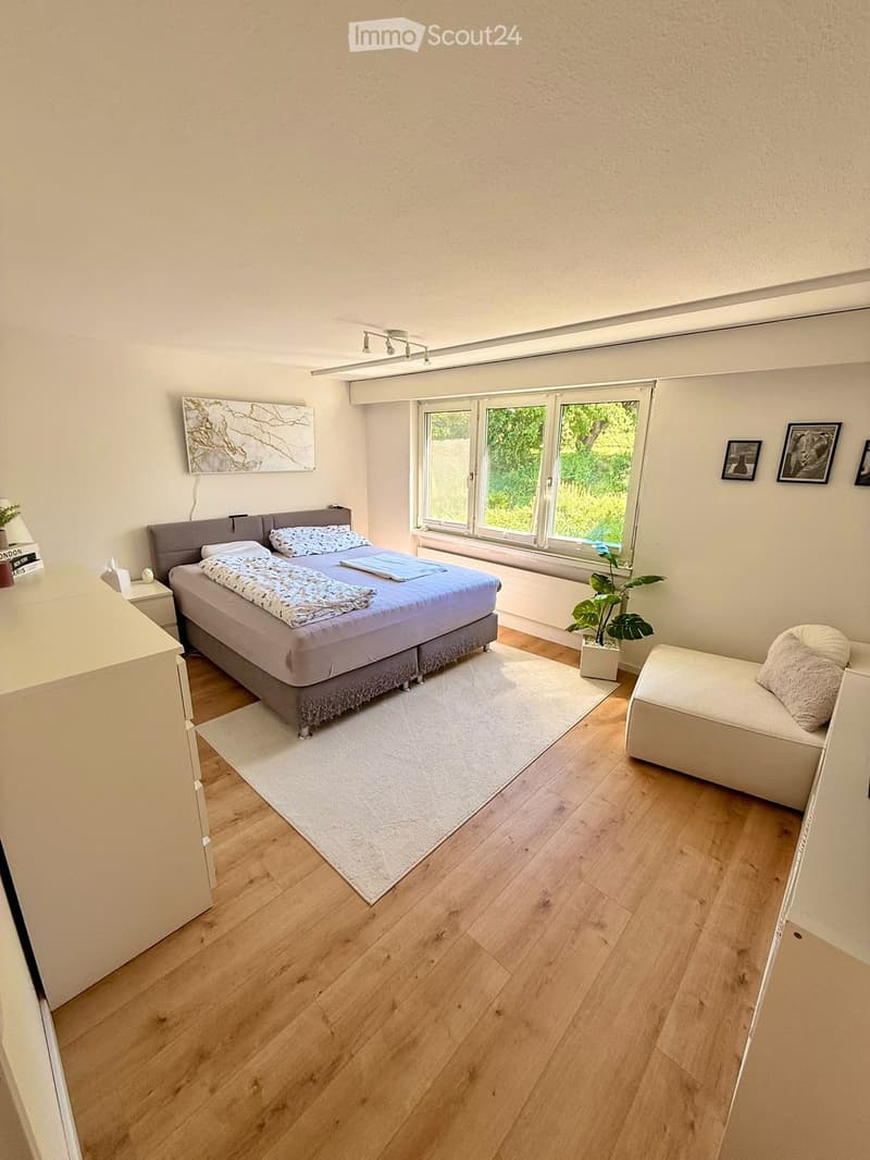 Modern bedroom with wooden flooring, large window, bed with pillows and blanket, nightstand, chair, and plant.