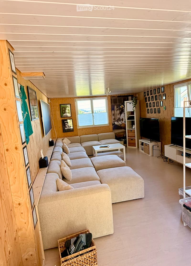 Cozy living room with a large sectional sofa, coffee table, ottoman, TV stand, and a woven basket on the floor. The room features wooden walls and large windows.