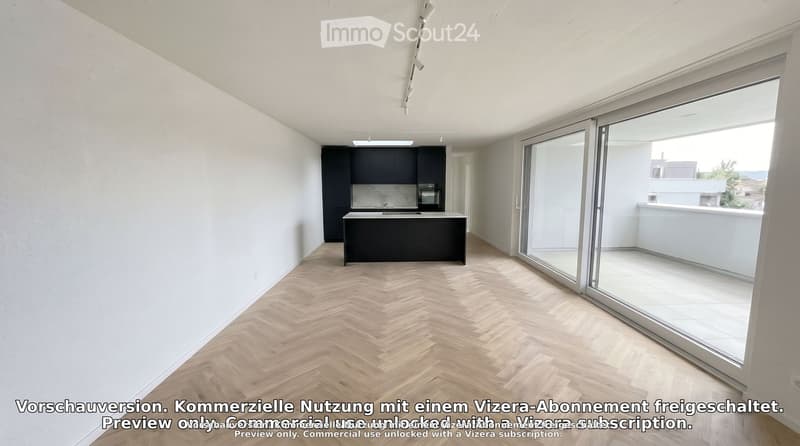 Open-plan space with a modern kitchen area featuring a black cabinet, island, sink, and oven, connected to a spacious living area with herringbone wooden flooring and large glass sliding doors leading to a balcony.