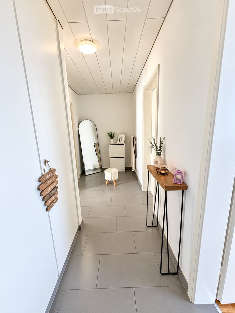 Modern hallway with white walls, grey tiled floor, and recessed lighting. Features a small wooden table with decorative items, a mirror, and a small stool.