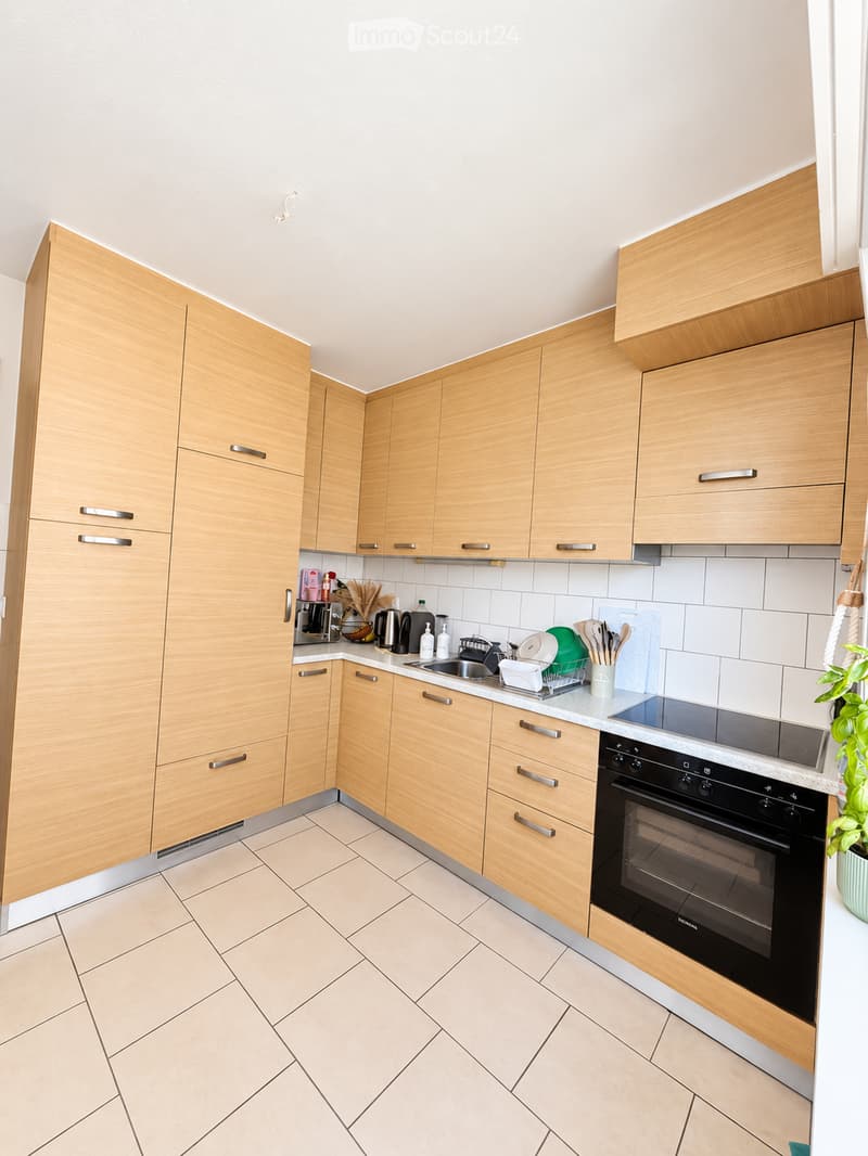 Modern kitchen with light wooden cabinets, stainless steel appliances, and a tiled floor. Features an oven, sink, dish rack, coffee maker, and various kitchen utensils.